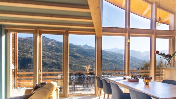 A spacious room with large windows offering mountain views, a dining area with chairs and a table, and wooden beams on the ceiling.