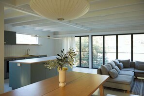A modern kitchen with a central island, a dining area with a wooden table, and a living room with a sofa. - My Malua,  Architectural Home by the Sea (Malua Bay)