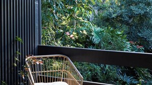 Eine Holzterrasse mit einem Korbstuhl, einem fensterartigen Metallrahmen und Blick auf einen üppigen Garten.