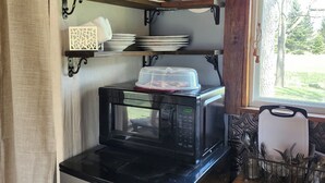 A compact kitchen with a stainless steel refrigerator, a microwave, and shelves holding dishes and cups. - Tiny House on Century Farmstead (Muskego)