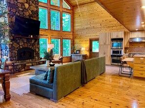 A spacious living area with a stone fireplace, a large window overlooking greenery, and a modern kitchen with stainless steel appliances. - Maxwell Reserve • Private Forest Retreat • Sleeps 14 (Tionesta)