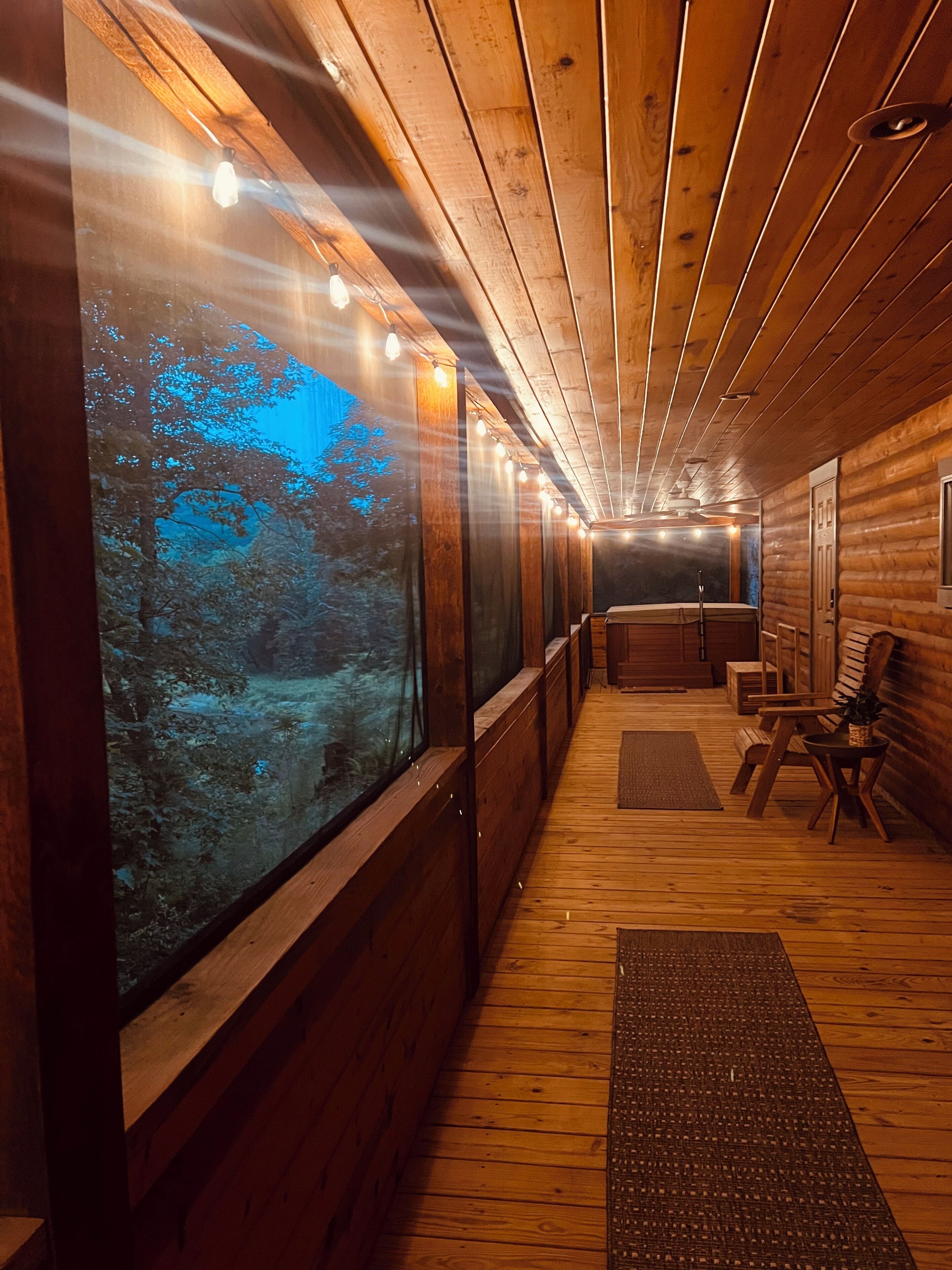A wooden cabin interior with a large window overlooking a forest, a seating area with a table, and a ceiling with exposed beams.