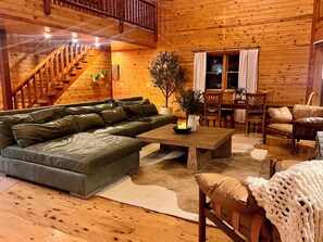 A cozy cabin interior with a wooden staircase, a sectional sofa, a dining table, and a large window with curtains. - Maxwell Reserve • Private Forest Retreat • Sleeps 14 (Tionesta)