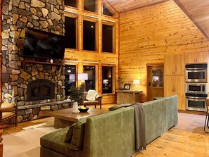 A cozy cabin interior with a stone fireplace, a television mounted on the wall, and a modern kitchen with built-in ovens. - Maxwell Reserve • Private Forest Retreat • Sleeps 14 (Tionesta)