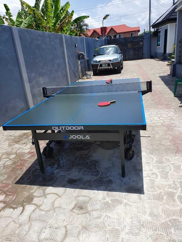 Una mesa de ping pong con estructura azul y la marca 'OUTDOOR', ubicada al aire libre sobre un suelo estampado, con un coche y un edificio al fondo.