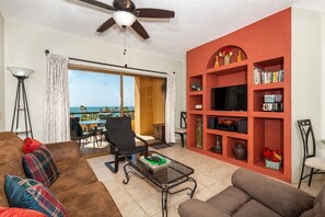 Ein Wohnzimmer mit Sofa, Sessel, Couchtisch und Bücherregal. Es gibt einen Deckenventilator, ein Fenster mit Vorhängen und einen Ausblick ins Freie.