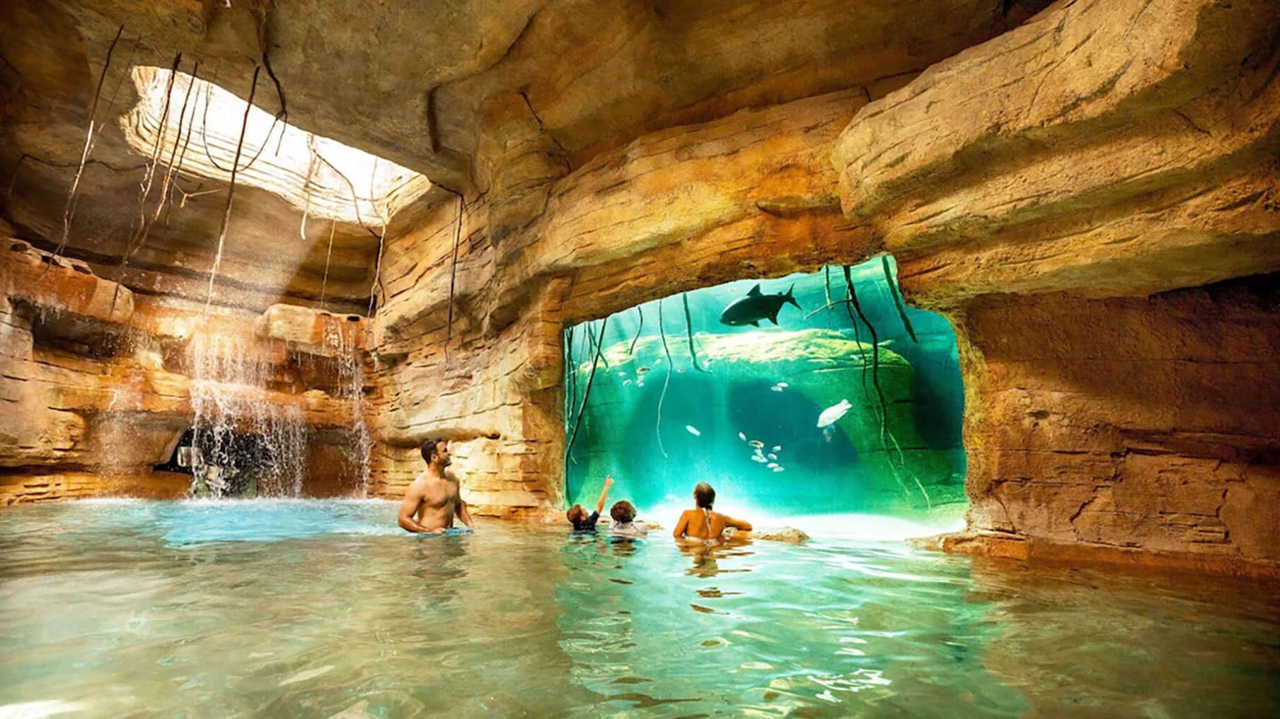 An indoor swimming pool with a large aquarium window, rocky walls, and a waterfall feature.