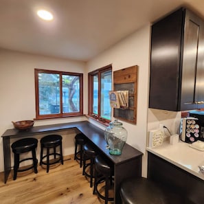 A kitchen with a bar area, wooden stools, a large window, and a cabinet with a coffee machine.
