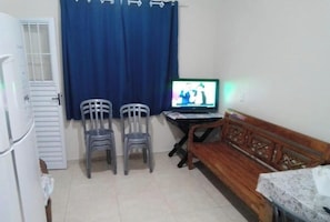 Een kleine kamer met een houten bankje, twee plastic stoelen, een tafel met een laptop en een raam met blauwe gordijnen.