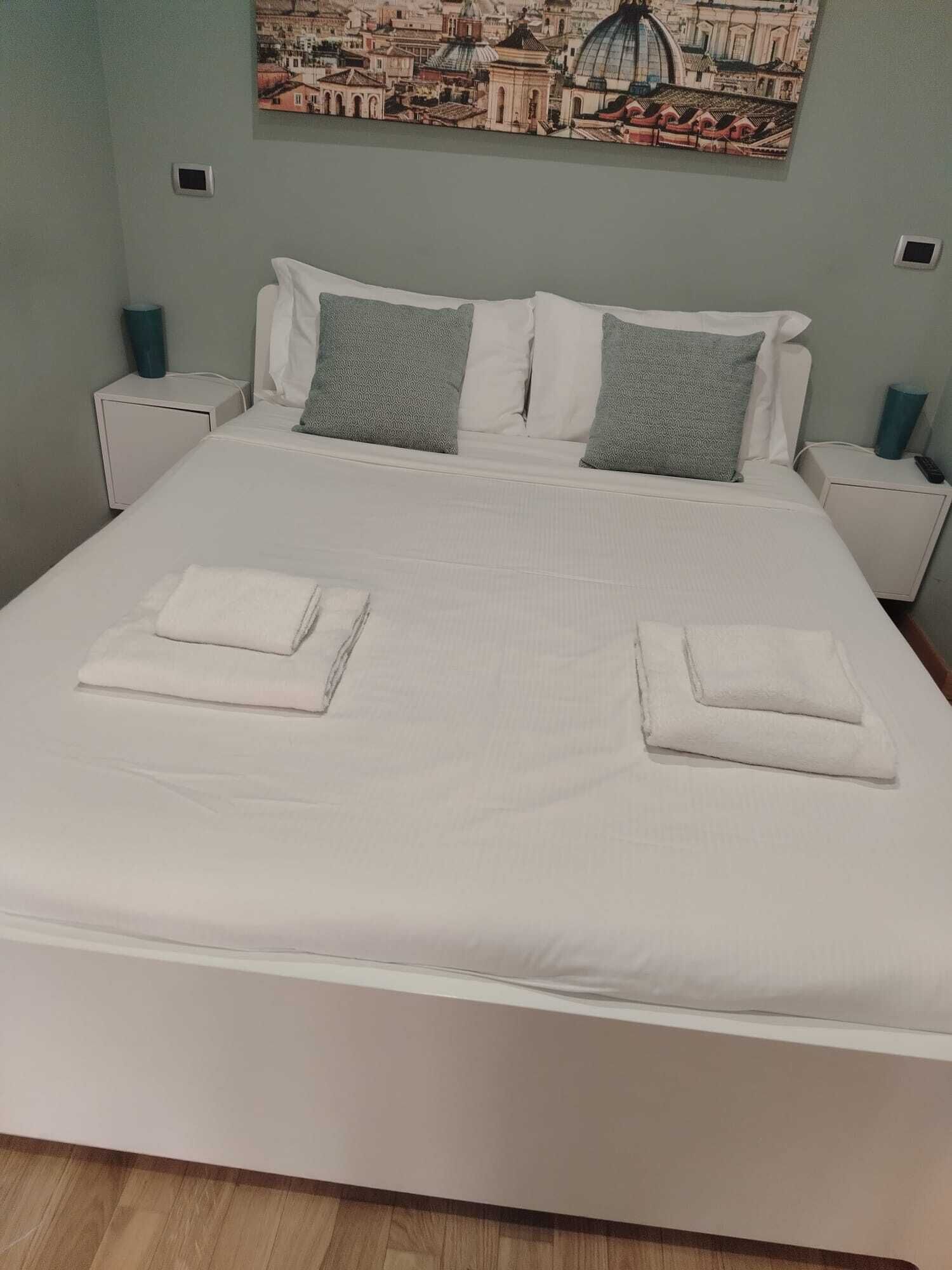 Un lit bien fait, avec du linge de lit blanc et des oreillers, un tableau représentant un paysage urbain accroché au mur, et une petite table de chevet avec une lampe.