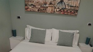 A neatly made bed with white linens and pillows, a cityscape painting on the wall, and a small bedside table with a lamp. - WONDERFUL VILLA BY THE SEA (Anzio)