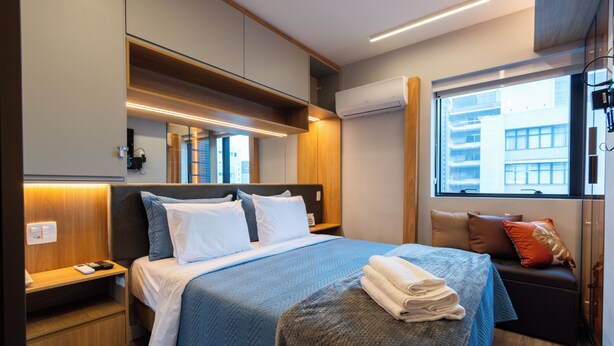 Una habitación de hotel moderna con una cama grande, una mesita de noche, un pequeño rincón de descanso y una ventana con vistas a edificios.