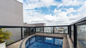 Una piscina en la azotea con vistas a la ciudad, rodeada de barandillas de cristal y plantas en macetas.