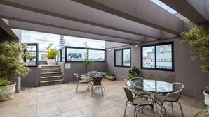 Una terraza en la azotea con una mesa y sillas de vidrio, plantas en macetas y vistas panorámicas de la ciudad.
