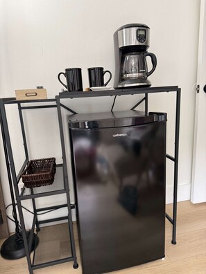 A black refrigerator with a coffee maker on top, a wicker basket, and a black metal shelf unit. - Private 1BR Studio • Patio • Free Parking • Near Beach (Lauderdale Lakes)