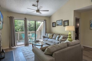 Ein Wohnzimmer mit einer Couch, einem Couchtisch und einem Deckenventilator. Ein Balkon bietet Blick auf Bäume.