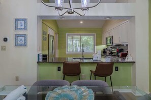 A modern kitchen with a large window, two bar stools, and a dining table with a glass top. - West Hyde Park 19D (Myrtle Beach)