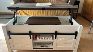Un deposito di auto bianco con ripiano superiore in legno, un libro intitolato 'SEQUENCE' e un cuscino su un divano sullo sfondo.