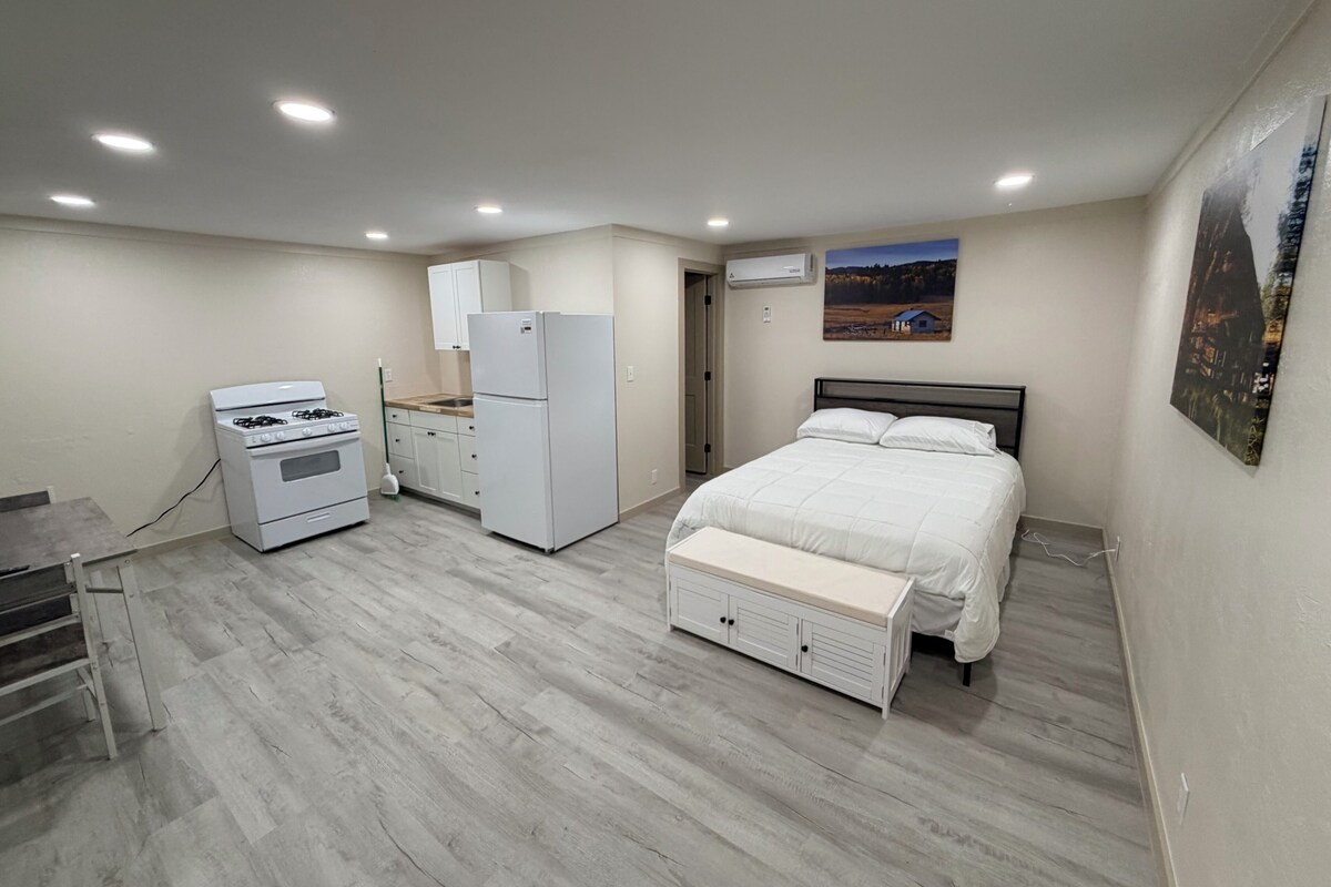 A bedroom with a bed, a nightstand, a dresser, a refrigerator, a stove, and a window with a view of a landscape.