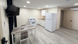 A compact kitchen with white appliances, a flat-screen TV mounted on the wall, and a small dining table.