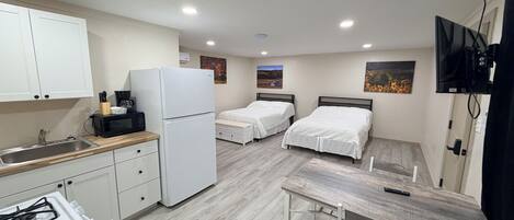 A compact kitchen with a refrigerator, stove, and microwave. There are two beds with white bedding and a TV mounted on the wall.