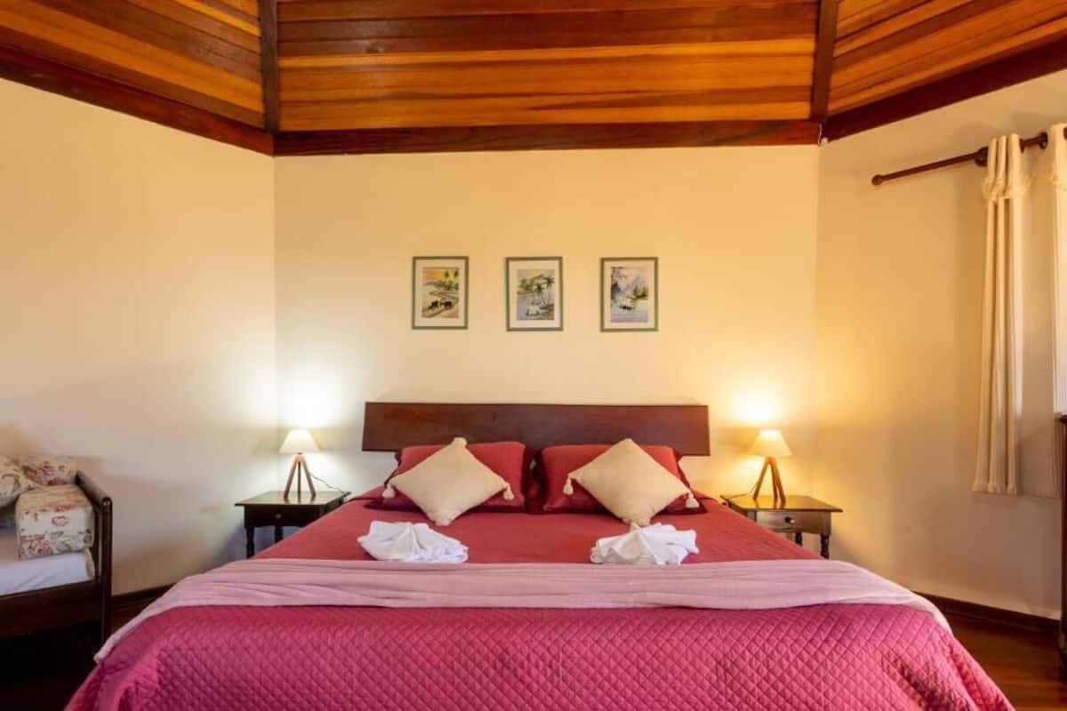 Habitación de hotel con techo de madera, una cama grande con ropa de cama roja, dos lámparas de noche y tres cuadros en la pared.