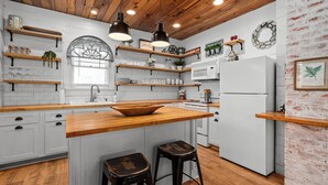 A kitchen with wooden countertops, white cabinets, and a central island with stools. There are hanging lights, shelves with kitchen items, and a refrigerator.