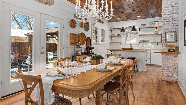 A dining area with a wooden table, white chairs, a chandelier, and a kitchen in the background.