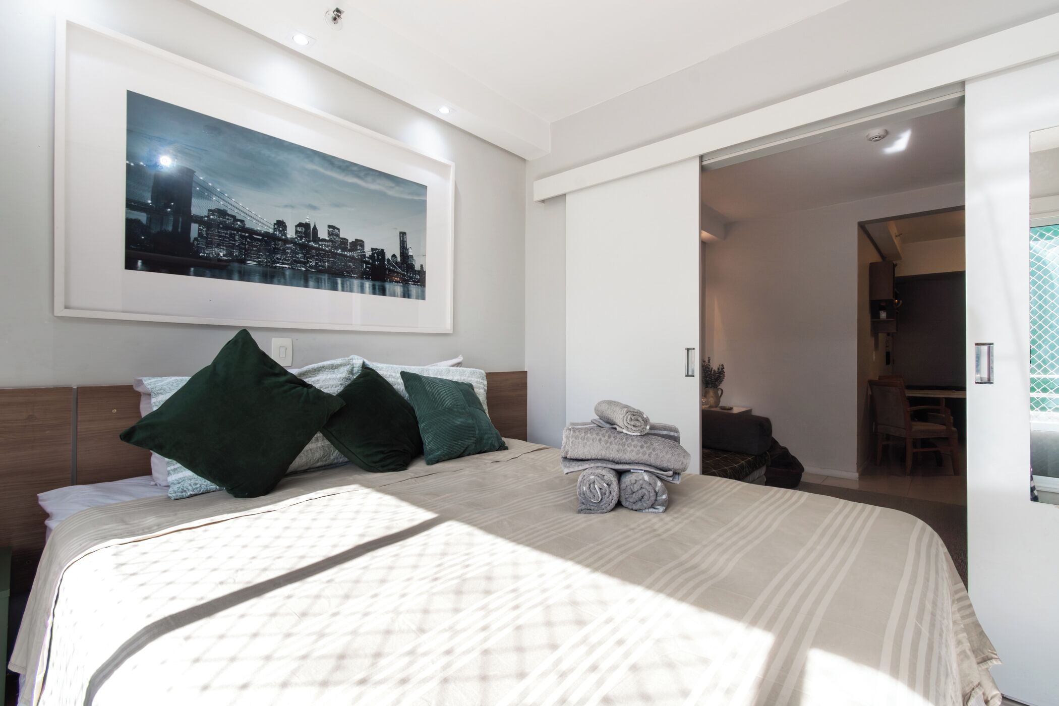A neatly made bed with a quilted cover, a framed cityscape picture on the wall, and a visible doorway leading to another room.