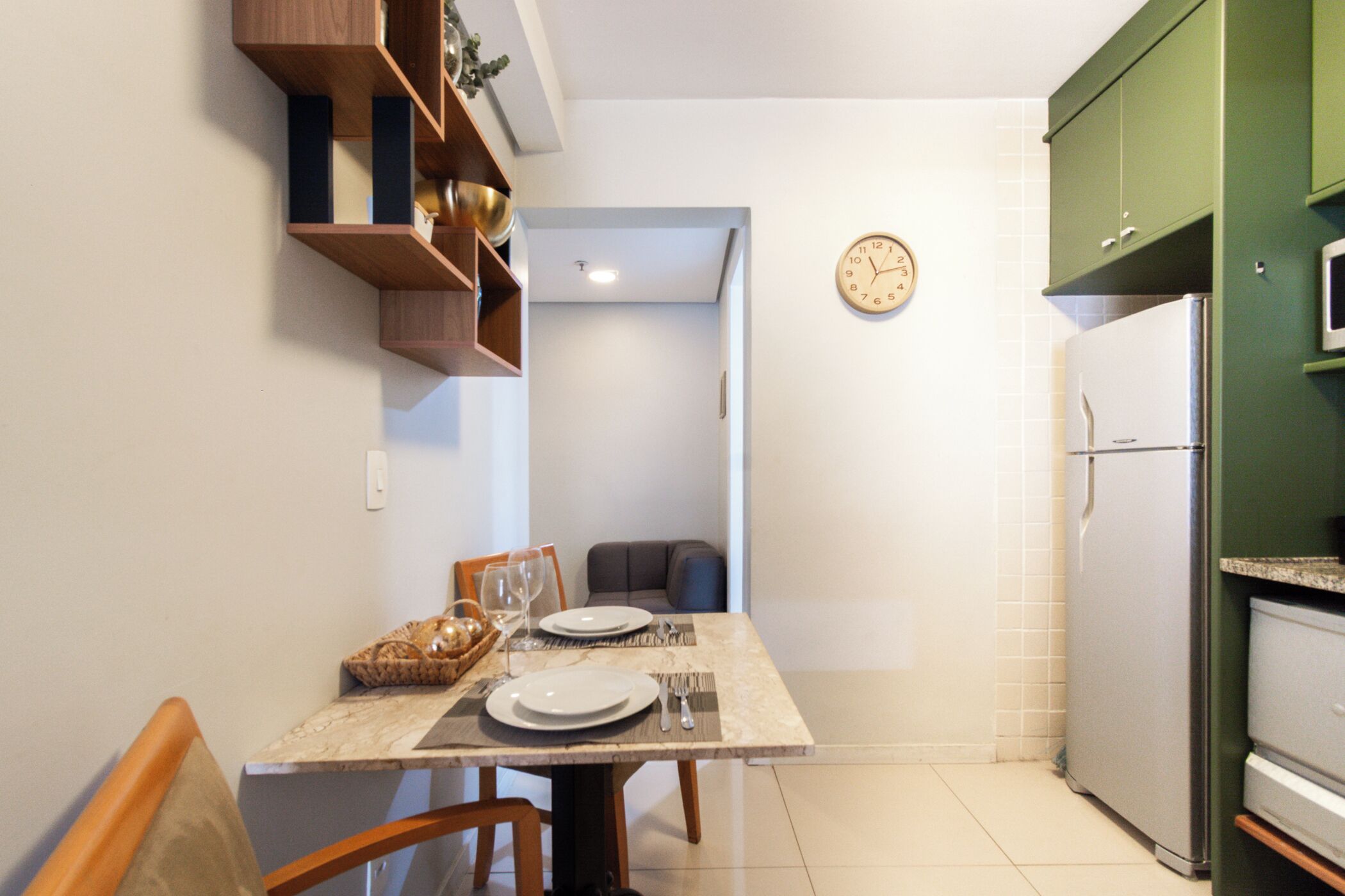 Een kleine keuken met een eettafel gedekt voor twee personen, een koelkast en een klok aan de muur.