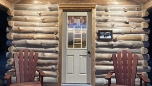 Une cabane en rondins avec une porte blanche, deux fauteuils Adirondack et une pancarte de bienvenue.