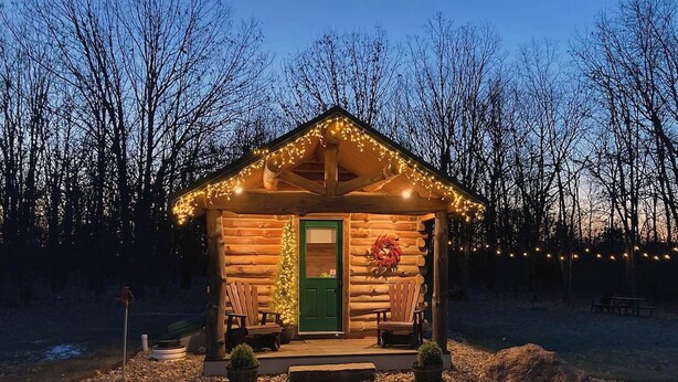 Une cabane en rondins avec une porte verte, deux chaises en bois à l’extérieur et des guirlandes lumineuses.