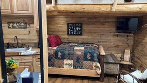 Une petite pièce comprenant un lit, une petite table, une chaise, un téléviseur et un mur en bois sur lequel est accroché un panneau.