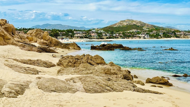 Ein Strand mit großen Felsen, klarem blauen Wasser und einer entfernten Hügellandschaft mit Gebäuden.
