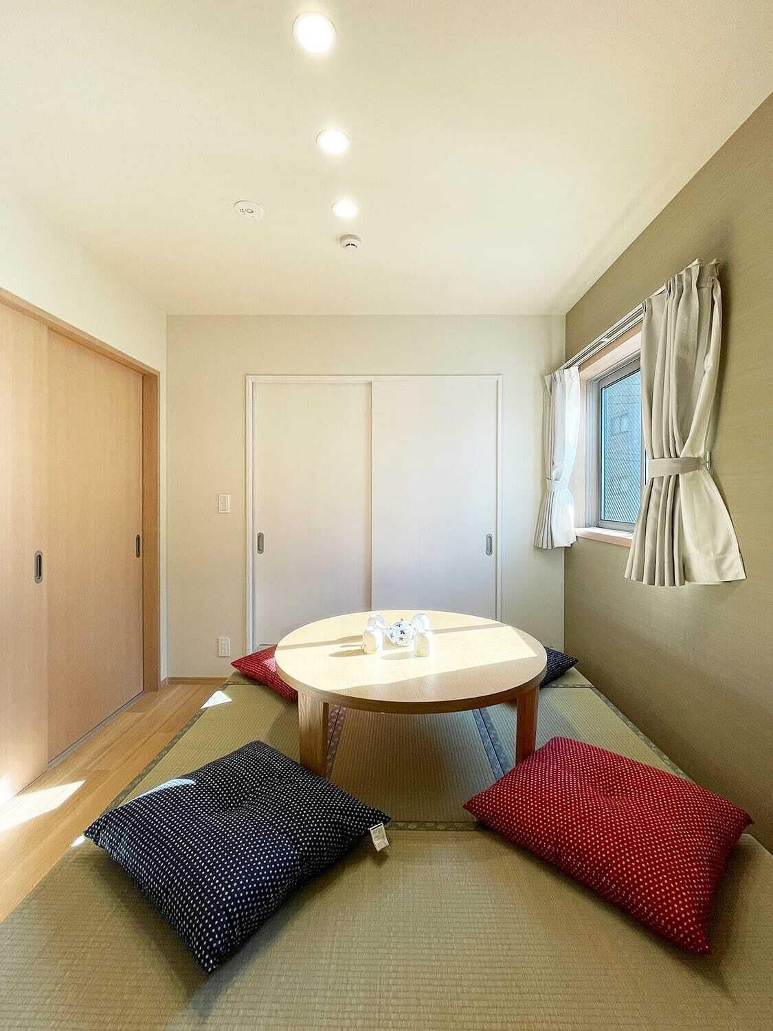 Ein Zimmer mit einem runden Tisch, zwei Kissen, einem Fenster mit Vorhängen und einer Schiebetür.