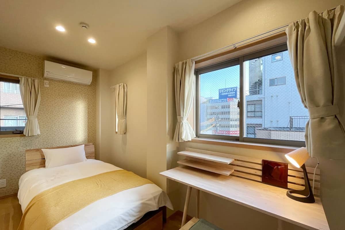 Una habitación de hotel con cama, escritorio con lámpara, ventana con cortinas y aire acondicionado en la pared.