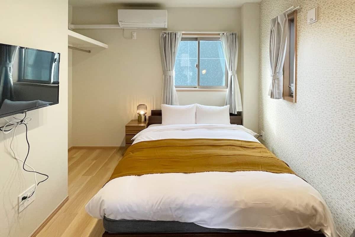 Un dormitorio con cama, televisor, aire acondicionado, ventana con cortinas y piso de madera.