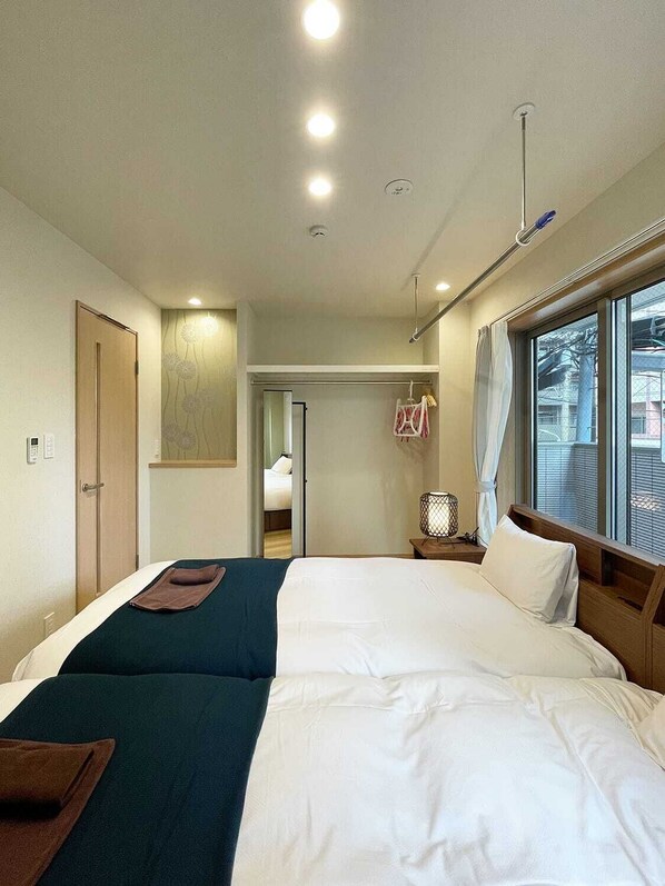A hotel room with two beds, a window with blinds, a bedside table with a lamp, and a door.