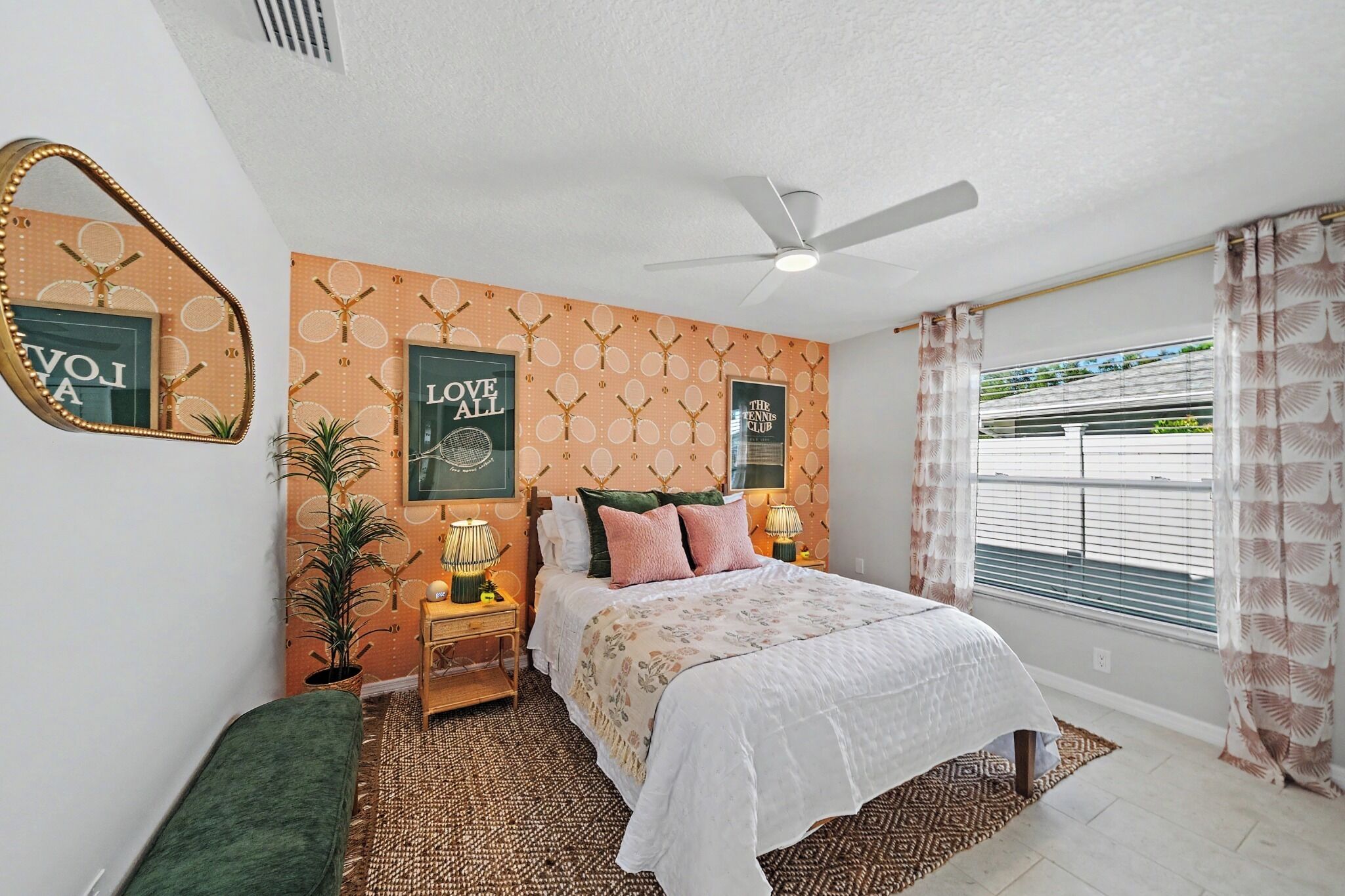 A bedroom with a bed, a ceiling fan, a window with blinds, a wall with tennis-themed decor, and a small table with a lamp.