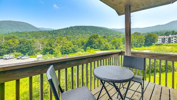 A wooden deck with two wicker chairs and a table, overlooking a lush green landscape with hills and a building in the distance.