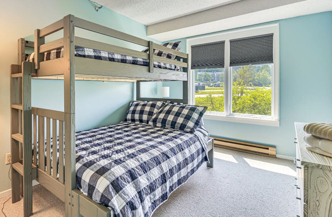 A bunk bed room with a checkered bedspread, a window with blinds, and a view of greenery outside.