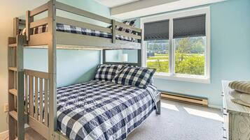 A bunk bed room with a checkered bedspread, a window with blinds, and a view of greenery outside.