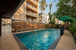 A swimming pool with a tiled deck, surrounded by a fence, and adjacent to a multi-story building with balconies. - Cozy 1bhk w Pool Gym Near Beach Kp-uf003 (Candolim)