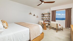 Quarto com cama, ventoinha no teto, janela com cortinas, cadeira e uma mesa pequena.