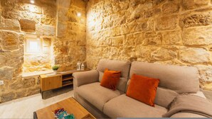 A stone-walled room with a beige sofa, a small table, and a window with a plant. - Charming House of Character in Victoria Gozo (Victoria)