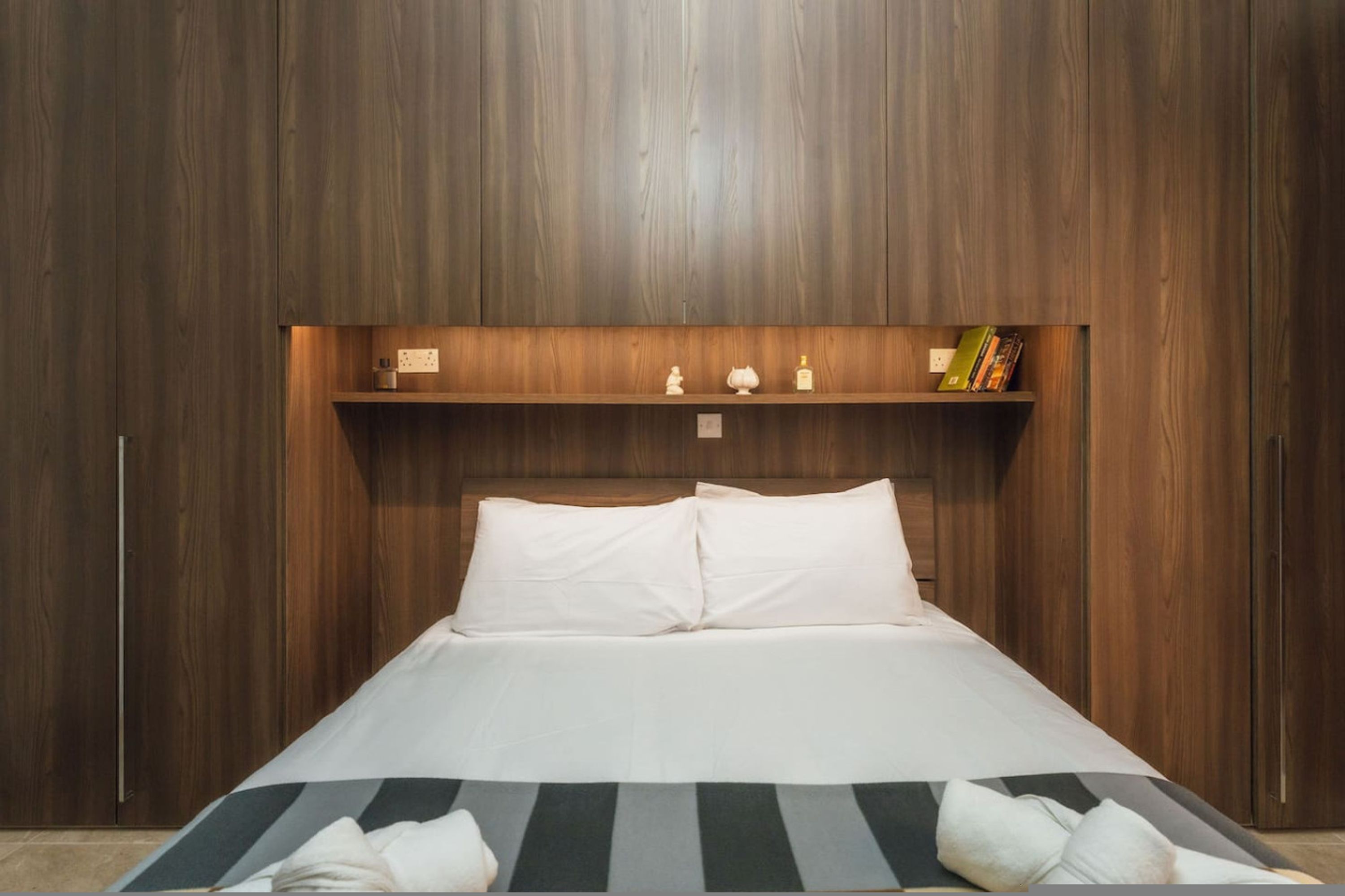 Une chambre à coucher avec une tête de lit en bois, un lit recouvert de draps blancs, une étagère avec des livres et des objets décoratifs, et un tapis à rayures.