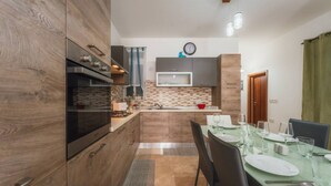 A modern kitchen with wooden cabinets, a built-in oven, and a dining area with a glass table set for four. - Marsalforn Apartment Walk to Beach Seaside Living (Zebbug)