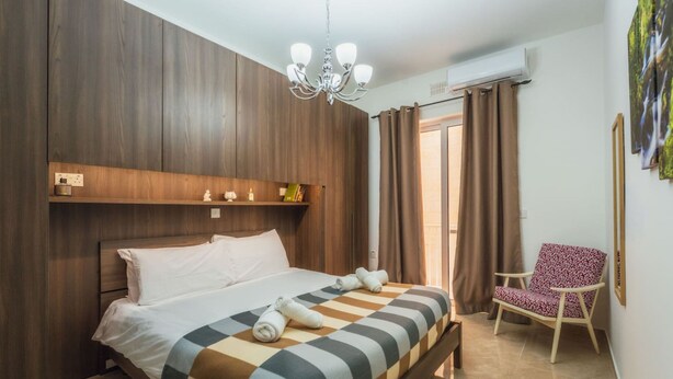 A bedroom with a bed, a chair, a chandelier, and a window with curtains. - Marsalforn Apartment Walk to Beach Seaside Living (Zebbug)