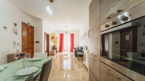 A modern kitchen with a large stove, dining area with a table set for two, and a living area with a sofa. - Marsalforn Apartment Walk to Beach Seaside Living (Zebbug)