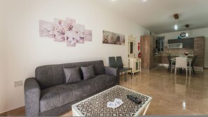 A living room with a grey sofa, a coffee table with a patterned design, and a dining area with a glass table and chairs. - Marsalforn Apartment Walk to Beach Seaside Living (Zebbug)